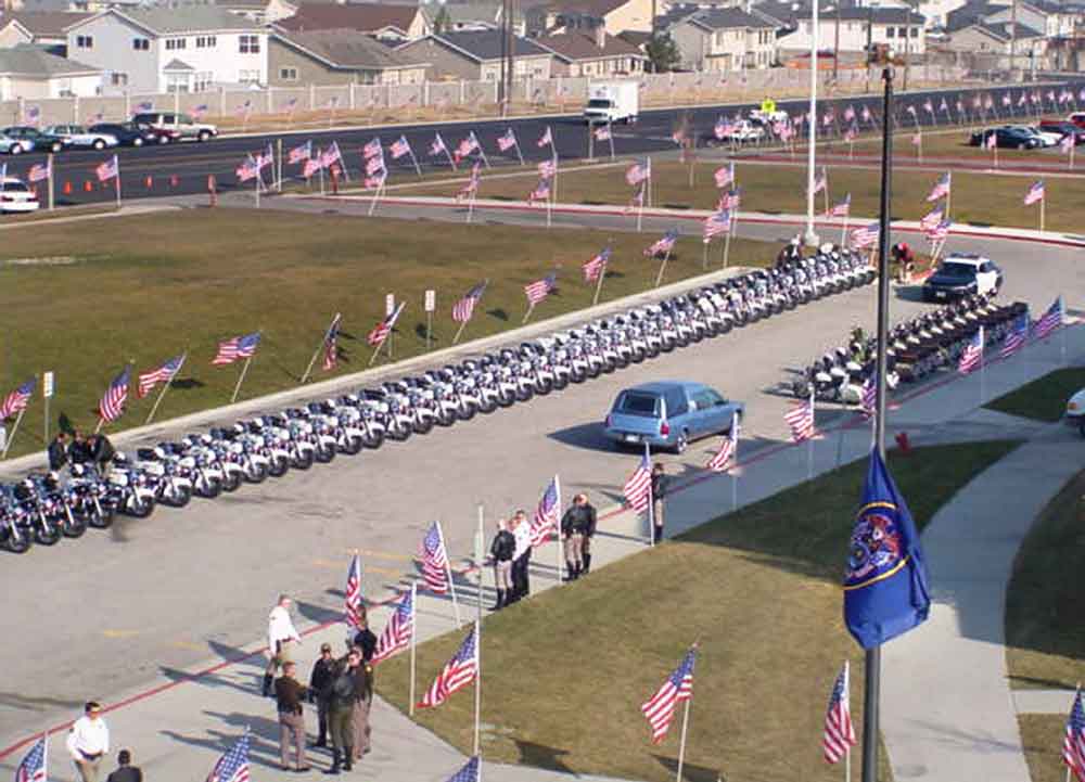 A blue hearse in front of rows of empty police motorcycles, The path is decorated with dozens of American flags.
