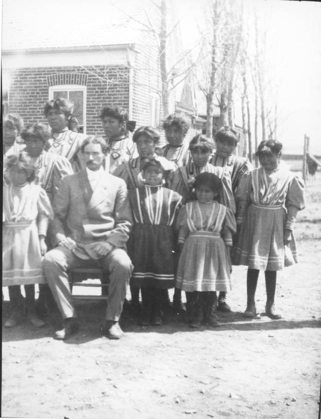 Historic photo of students in and an adult man sitting on a bench