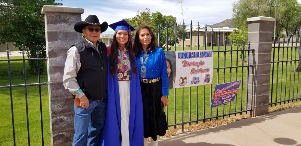 A woman in a cap and gown smiles with her parents in front of a fence